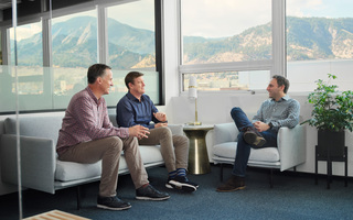 Three men sitting on grey couches in front of a window