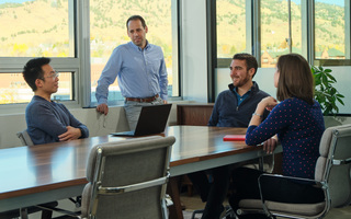 Three men and one woman around a conference table with a window in the background.